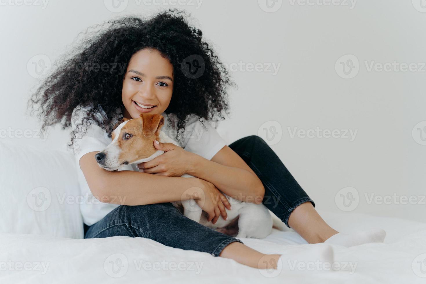 Photo of lovely young female pet owner poses in clean white bedroom, hugs dog, plays with best friend, wears casual clothing, has cheerful expression. People, animals, love, friendship concept
