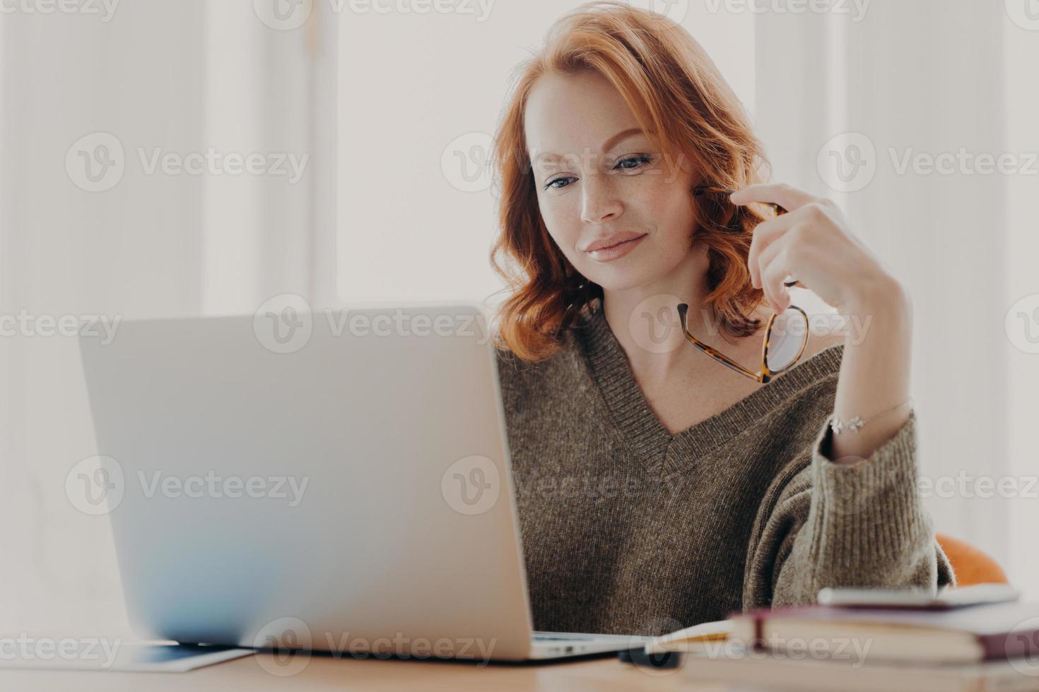 Concentrated female student reads information and checks data, focused at laptop screen, uses wireless internet connection, prepares for exam, poses at workplace, holds eyewear for vision correction photo