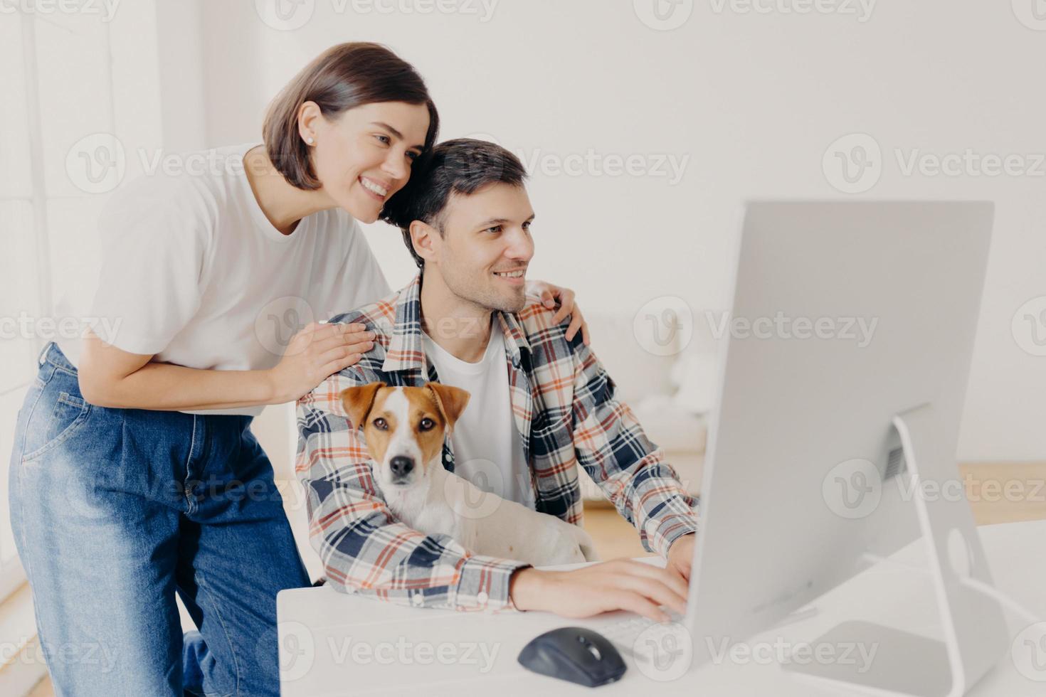 Image of woman touches gently husbands shoulders, look positively in monitor of computer, surf internet, buy furniture online for their new apartment. Happy man works at modern device with dog photo