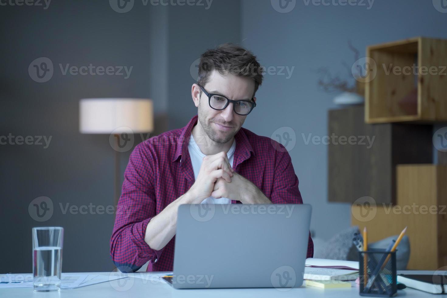 Pensive young German man home office employee sits at desk at workplace operates online on laptop photo
