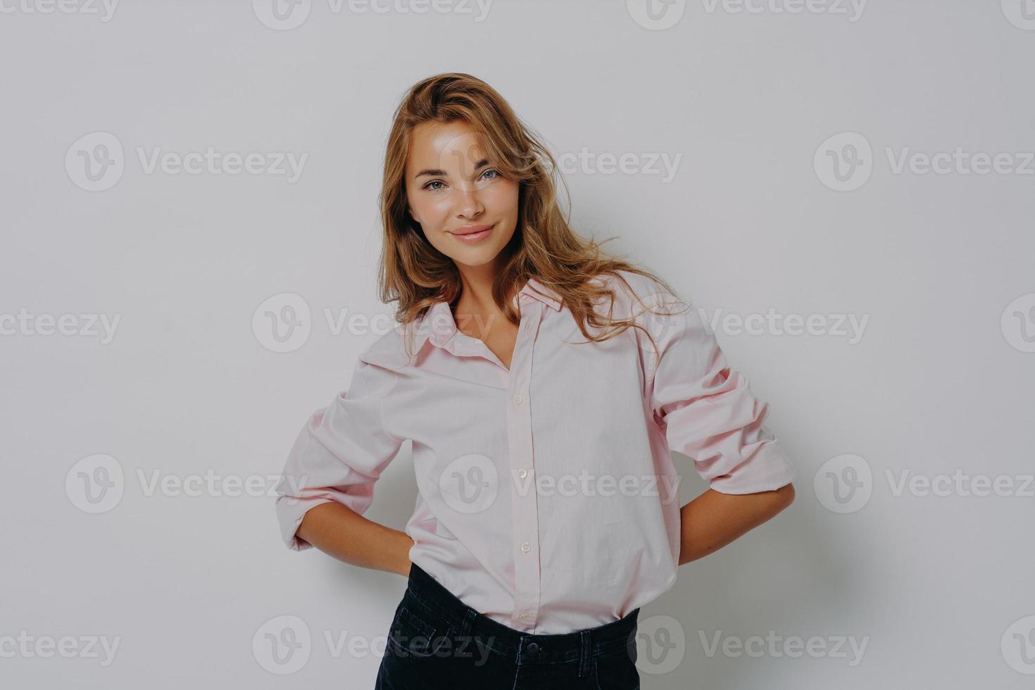 Elegant attractive female model in casual shirt and jeans standing with folded hands behind her back photo