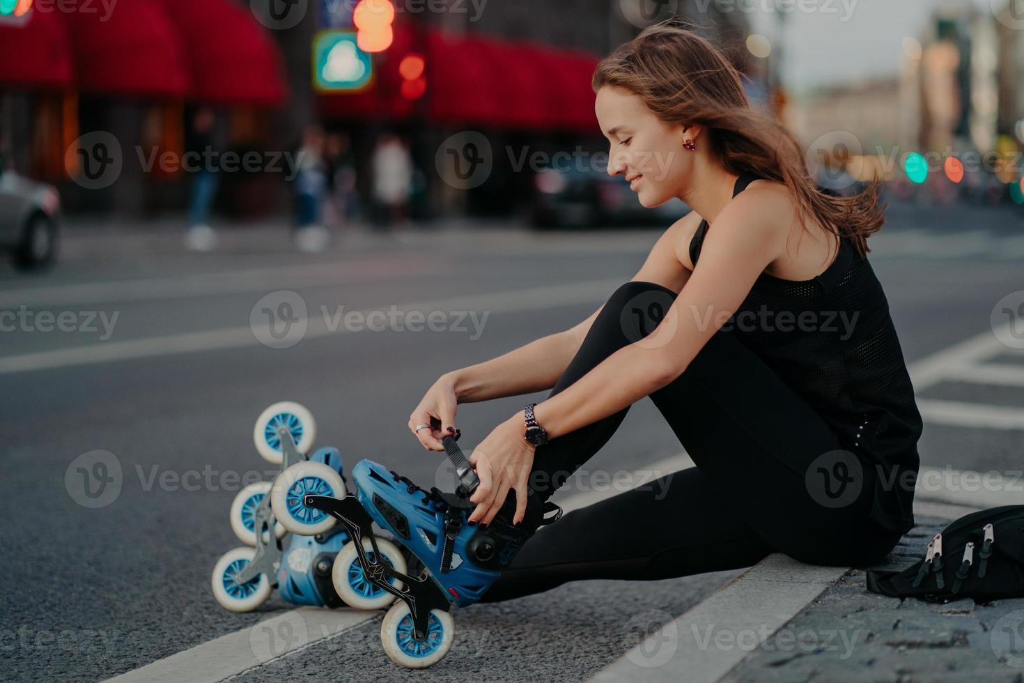 Outdoor shot of active woman laces rollerblades prepares for ride sits on road against busy city background dressed in black activewear enjoys rollerskating. Sporty lifestyle and recreation concept photo