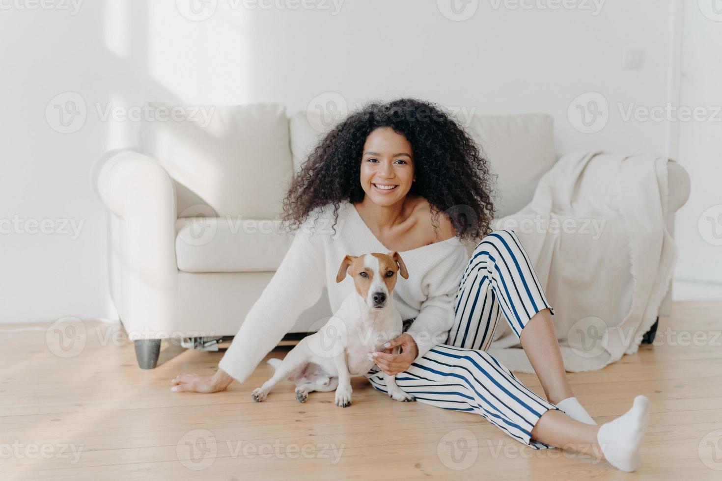 Relaxed young African American woman sits on floor in empty room near sofa with pedigree dog, enjoys sweet moment at home, move in new apartment to live. Lady rests with puppy in living room photo