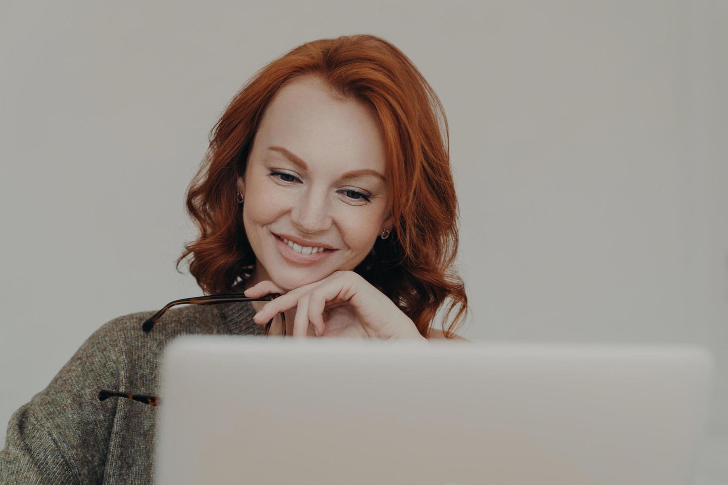 una estudiante exitosa con cabello pelirrojo y una sonrisa agradable ve un video tutorial interesante o un seminario web, usa una computadora portátil para el aprendizaje electrónico, lee información en línea, mantiene la mano debajo de la barbilla foto