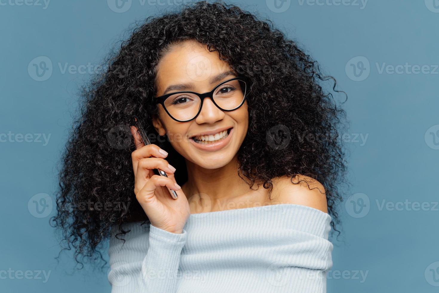 retrato de mujer afroamericana satisfecha habla por teléfono móvil, disfruta de una buena conversación, cuenta noticias a los mejores amigos, sonríe ampliamente mientras escucha una historia divertida, aislada sobre fondo azul. tecnología foto