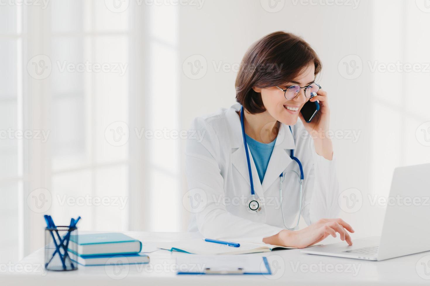 Online consultation concept. Cheerful female medical worker talks via cellular, searches useful information in internet, works on laptop computer, sits at desktop with notebooks, clipboard, pens photo