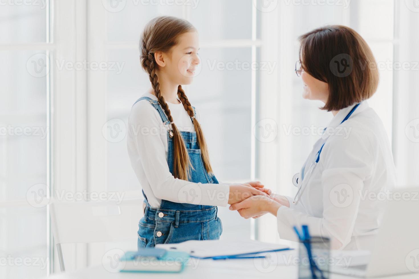 Female doctor shakes hands with girl child patient, talks supportive words, cares about kid during professional consultation, ready always to help with good treatment. Medical insurance concept photo