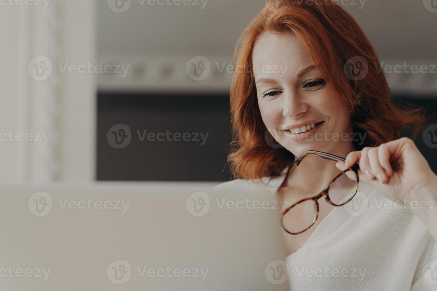 Photo of good looking cheerful redhead woman reads information on website in internet, works on laptop computer, holds transparent glasses, works on freelance project from home, shares content online