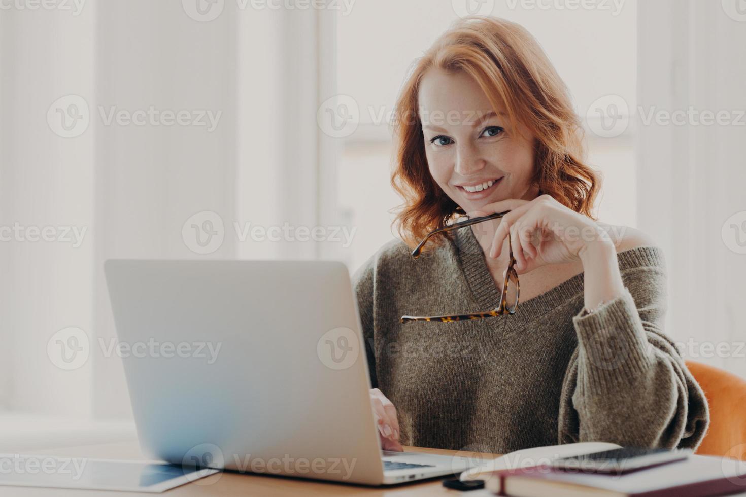 mujer pelirroja confiada y alegre trabaja en una computadora portátil moderna, posa en un espacio de trabajo compartido, usa suéter, trabaja de forma remota con una aplicación en el dispositivo, sonríe agradablemente, sostiene espectáculos, disfruta del trabajo a distancia foto