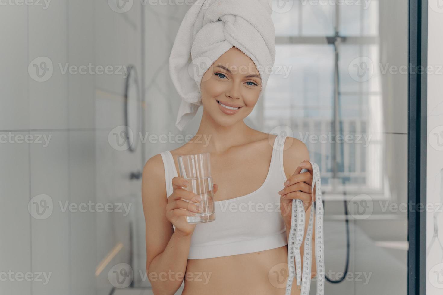 mujer caucásica sonriente y atractiva con un vaso de agua en una mano y cinta métrica en la otra foto