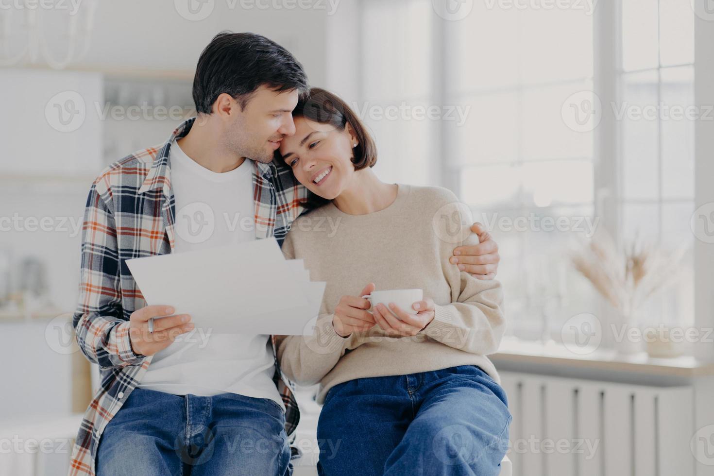 Caring husband embraces wife with love, have glad expressions, holds paper documents, discuss banking procedures, study bills, drink tea or coffee, pose indoor, enjoy calm domestic atmosphere photo