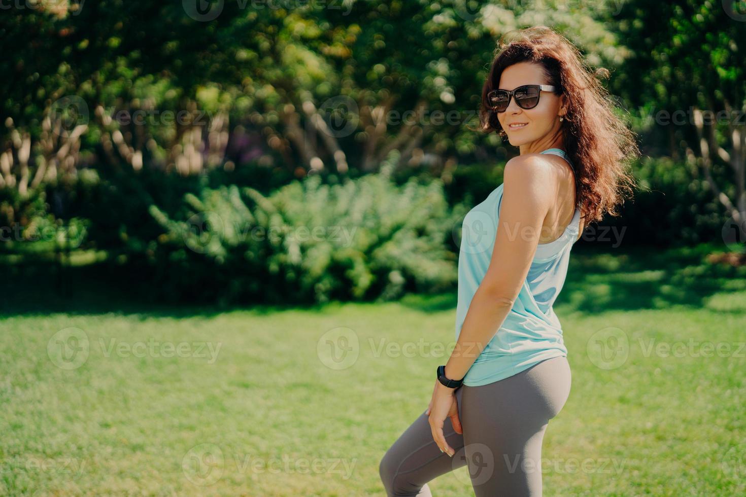 la mujer europea morena complacida en ropa activa posa de lado usa sunglases smartwatch tiene fitness al aire libre descansa después de trotar sonríe agradablemente. salud cuidado del cuerpo deporte y ejercicio concepto foto