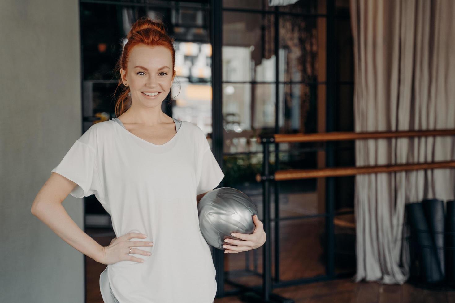 Positive fitness instructor with ginger hair posing with small silver fitball in hand photo