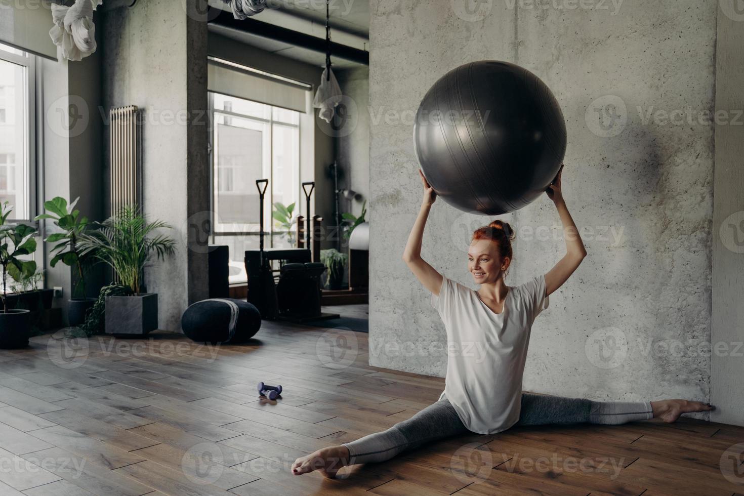 Beautiful fitness woman in split position holding exercise ball above head and enjoying pilates workout photo