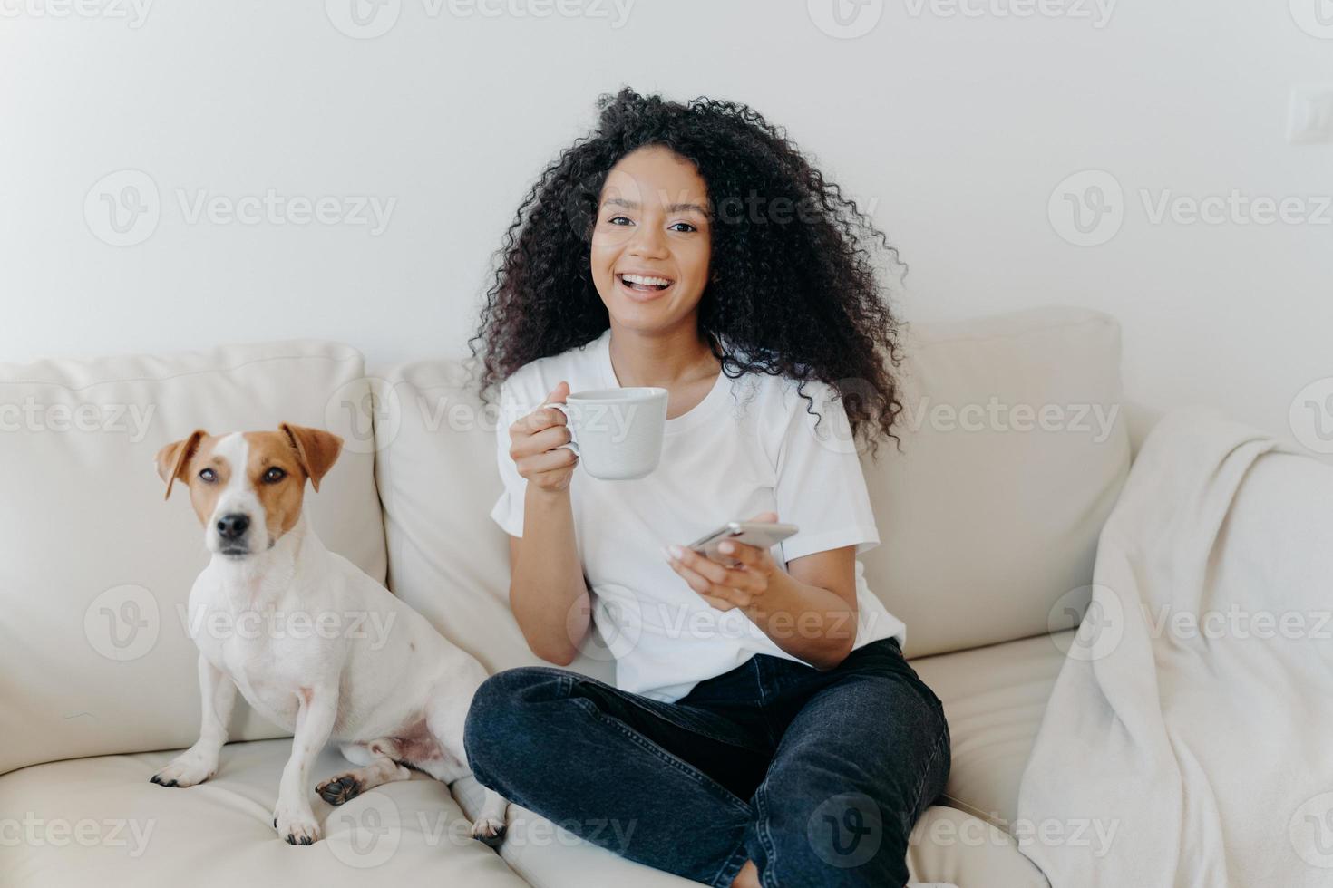 Delighted dark skinned woman poses in modern apartment, sits on comfortable sofa with pet, drinks coffee, uses mobile phone for online communication, being in good mood, flipping news, uses app photo