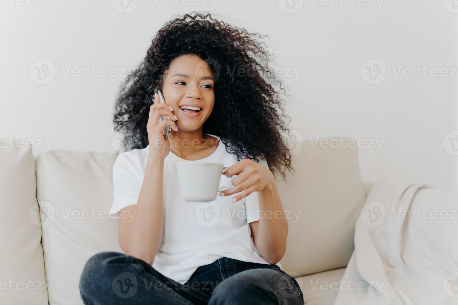 niña milenaria sonriente con cabello afro, disfruta de una conversación por teléfono celular con una bebida caliente en una taza, tiene una piel sana, se viste informalmente, se sienta en un sofá blanco en casa en un apartamento moderno, ordena la entrega foto