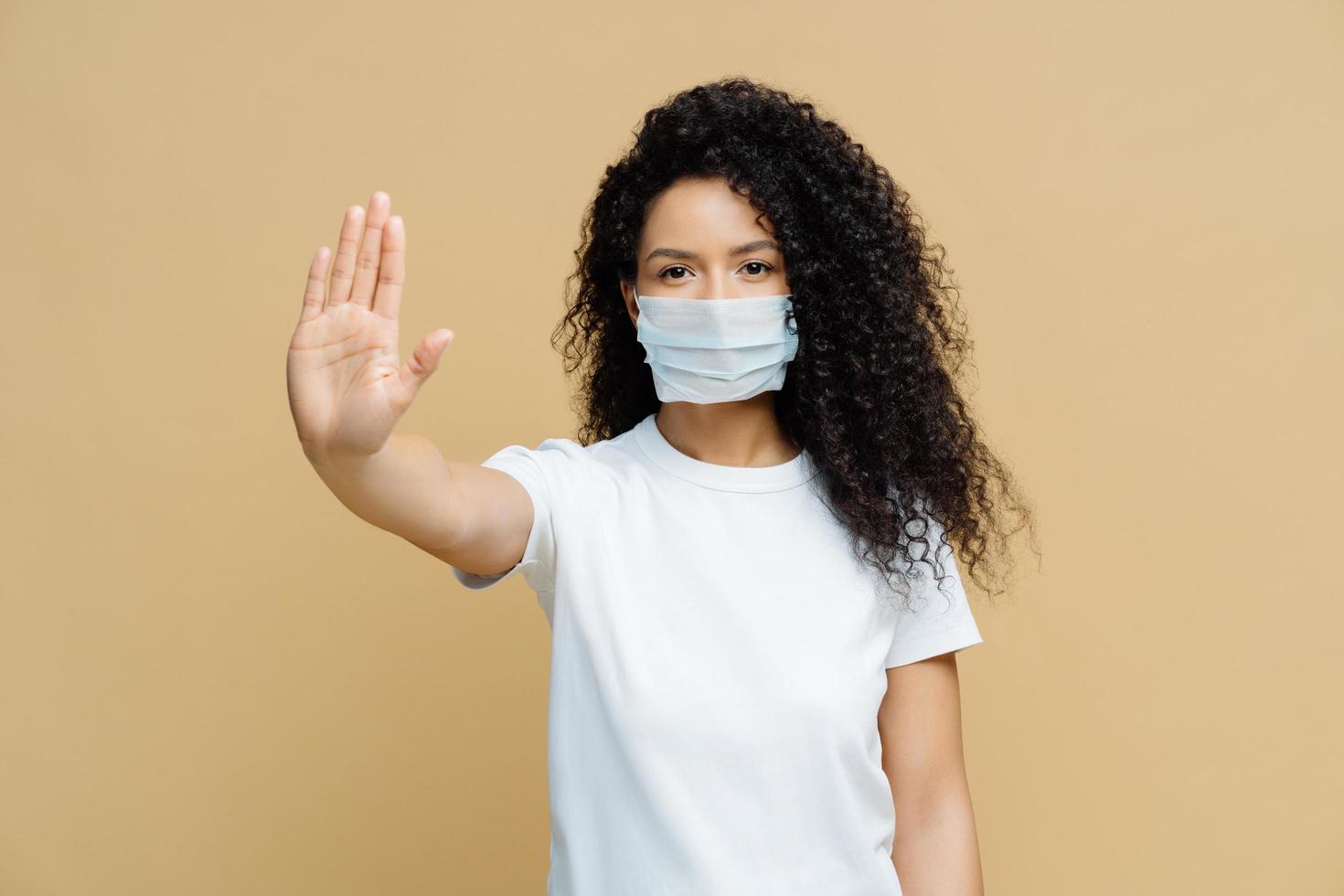 Serious dark skinned woman makes stop gesture, pulls palm towards camera, wears medical flu mask, asks stay at home not to spread coronavirus disease, prevents virus, dressed in white t shirt. photo