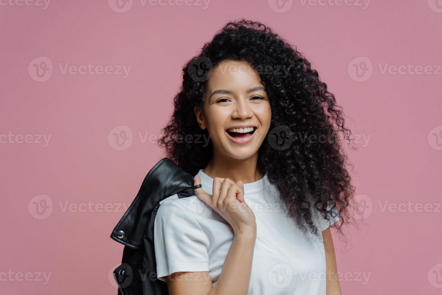 Portrait of positive African American woman smiles toothily, being in good mood after walk in park, dressed in white t shirt holds leather jacket on shoulder isolated on pink background. People, style photo