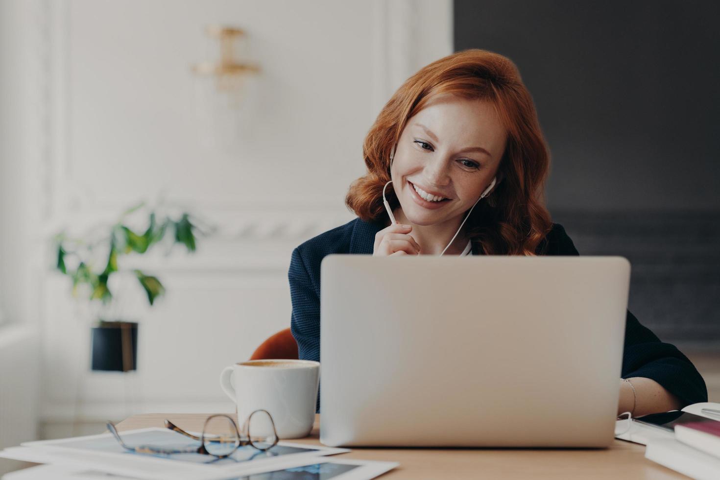 Positive ginger businesswoman works remotely from home during quarantine, has video conference with colleagues, makes online call, uses wireless internet and modern laptop computer with earphones photo