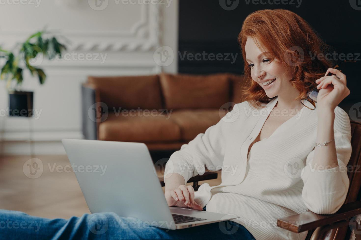 Skilled positive female student creats article for homework, sits in coworking space, has pleased expression, works on laptop computer, pose over home interior, prepares for writing course work photo