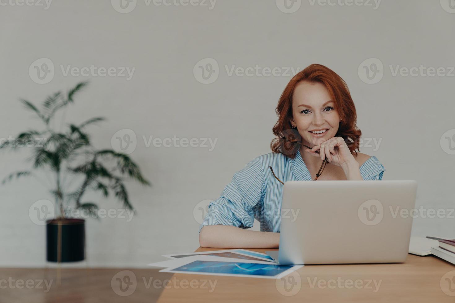 Cheerful foxy female entrepreneur chats online with business partners, works on startup project, poses at workplace, surrounded with paper documents, white wall and potted flower in background photo