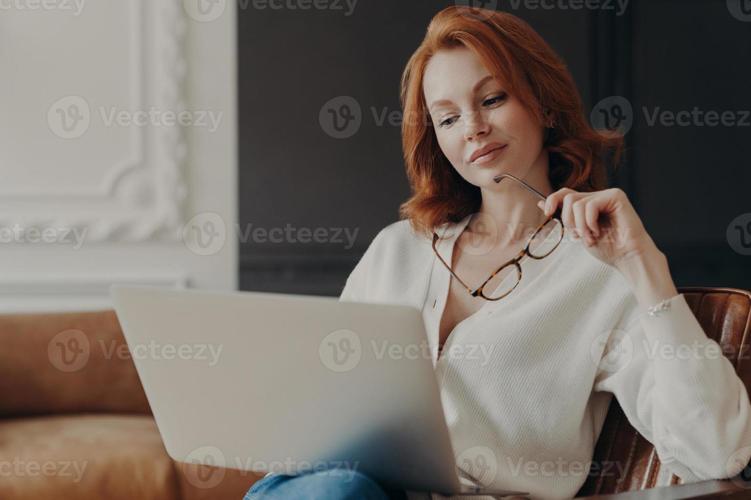 Concentrated serious redhead woman makes remote job indoors, sits in comfortable armchair, holds transparent glasses, reads publication on internet website, uses home wifi. Working time, technology photo