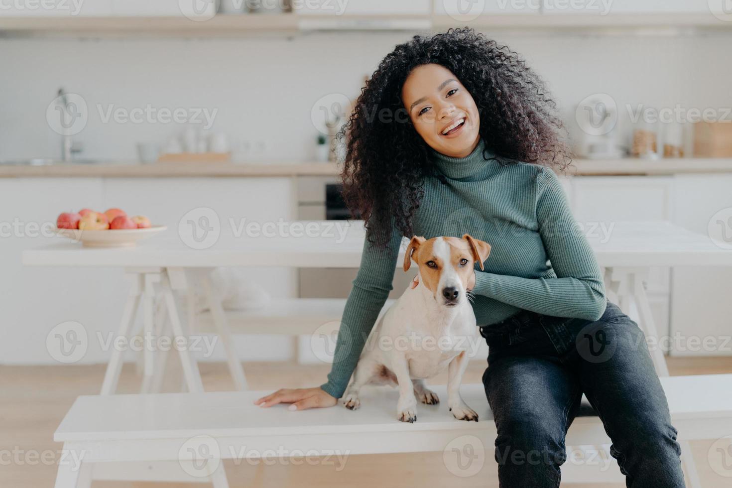 la dueña de una mascota expresa su amor por el perro, vestida con un cuello de tortuga informal y jeans, se sienta en un banco blanco contra el acogedor interior de la cocina, pasa tiempo libre en casa, sonríe ampliamente de felices emociones foto