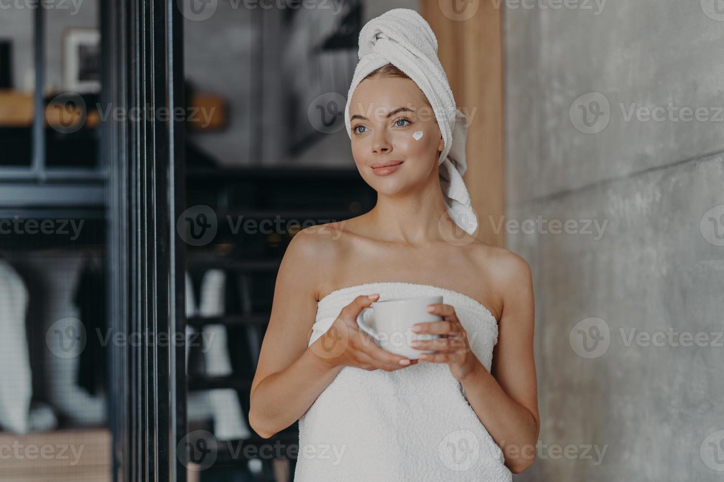 Thoughtful lovely young female model with cream on face, wrapped in towel, concentrated somewhere aside, holds mug of tea, undergoes beauty treatments at home, stands indoor. Body care concept photo