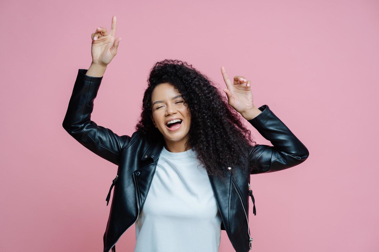 Carefree beautiful Afro American woman dances with arms raised, moves with rhythm of music on disco party, wears white t shirt and leather jacket, closes eyes with pleasure, keeps mouth opened photo