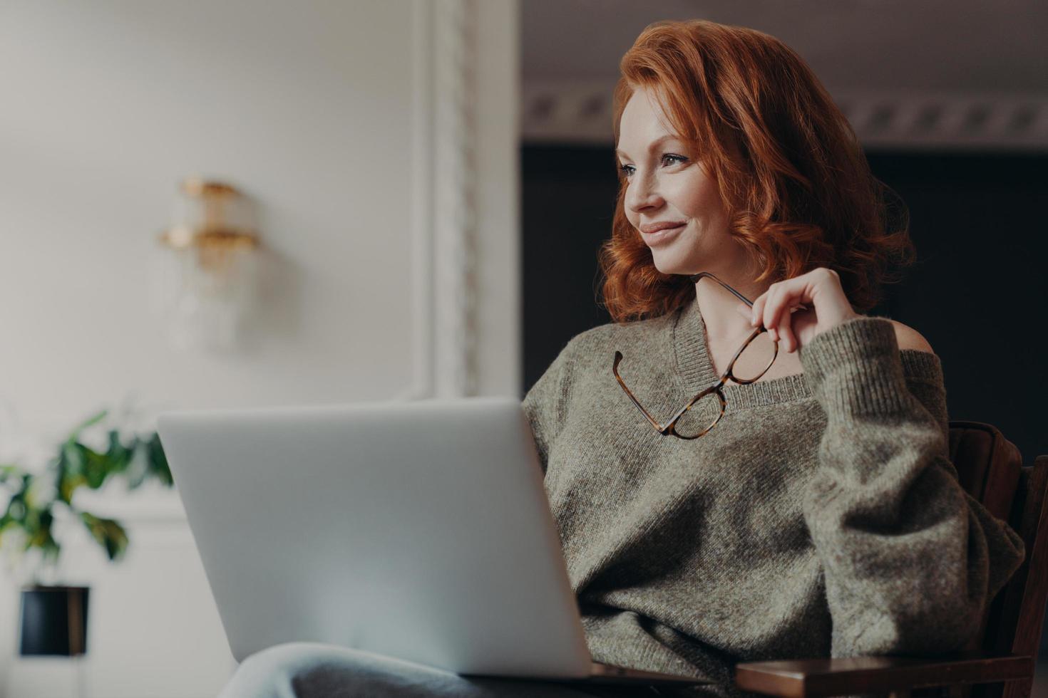 Experienced redhead female entrepreneur makes plans, looks aside with dreamy expression, uses modern technology, holds spectacles, wears casual jumper, enjoys distance education and e learning photo