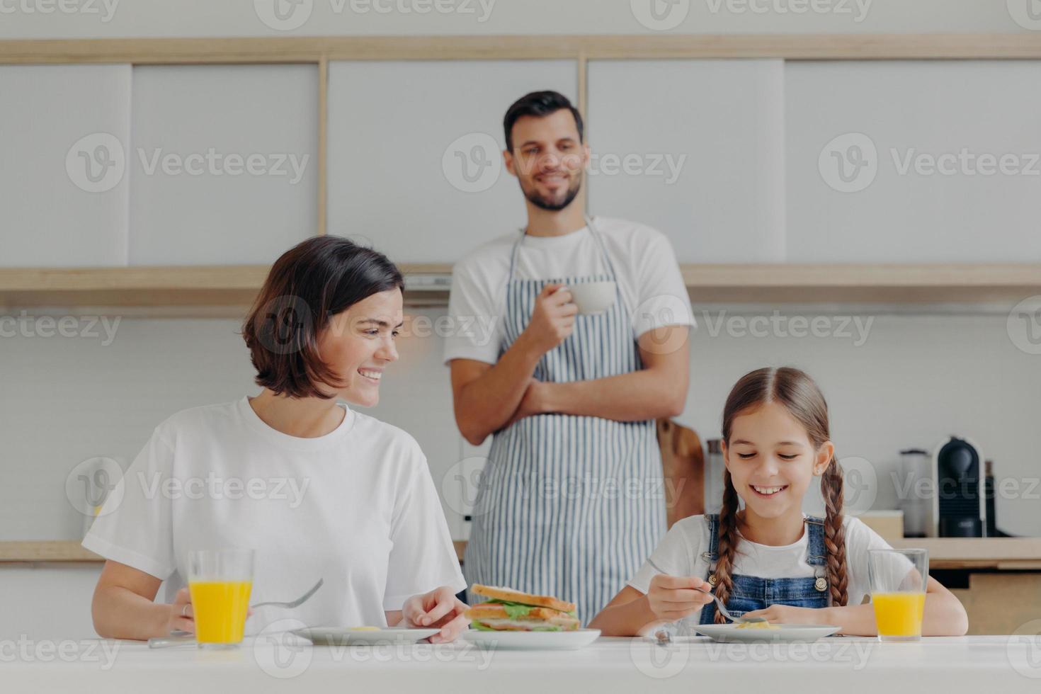 Happy mother talks to daughter while have breakfast. Father stands behind, prepared delicious dish for family. Friendly family members meet at kitchen during weekend, enjoy nice conversation photo