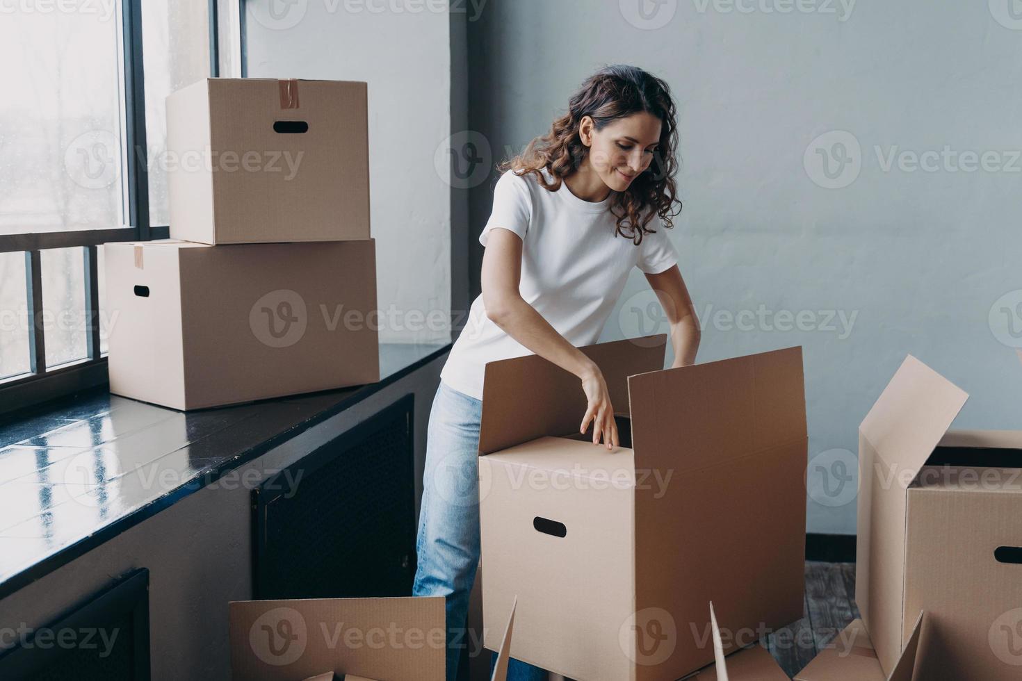 Happy young european woman unpacking boxes. Girl near window opening cardboard box and smiling. photo