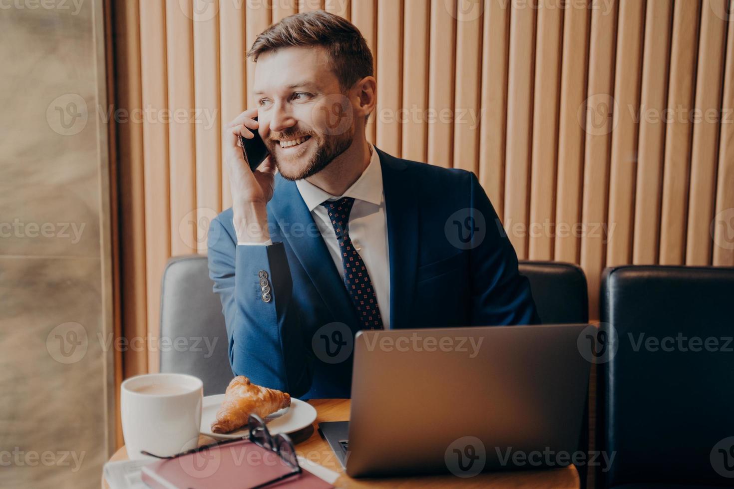 un apuesto hombre de negocios sin afeitar sonriente con traje formal hablando por celular en un café foto