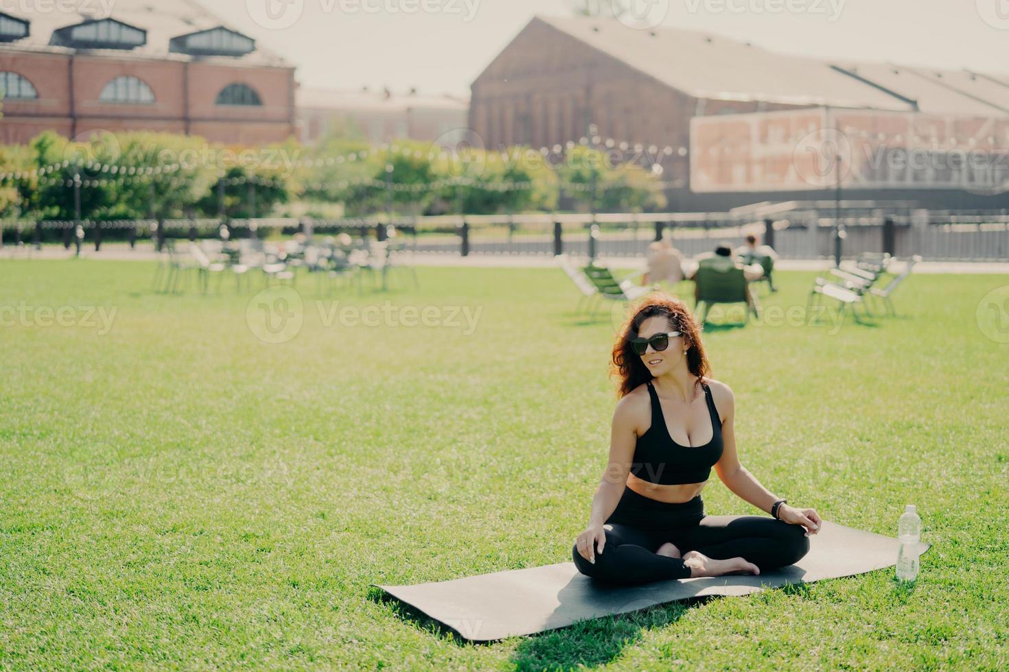 Positive dark haired woman sits crossed legs with bare feet on karemat dressed in activewear looks gladfully aside enjoys sunny day wears sunglasses practices yoga outdoors. Sporty lifestyle concept photo