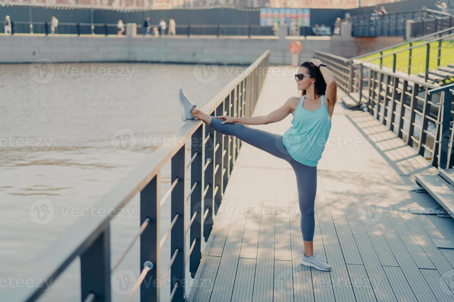 mujer joven en forma con cabello oscuro estira las piernas en la valla se calienta antes de trotar usa gafas de sol camiseta polainas y zapatillas de deporte se prepara para el entrenamiento cardiovascular plantea al aire libre estar en buena forma física foto