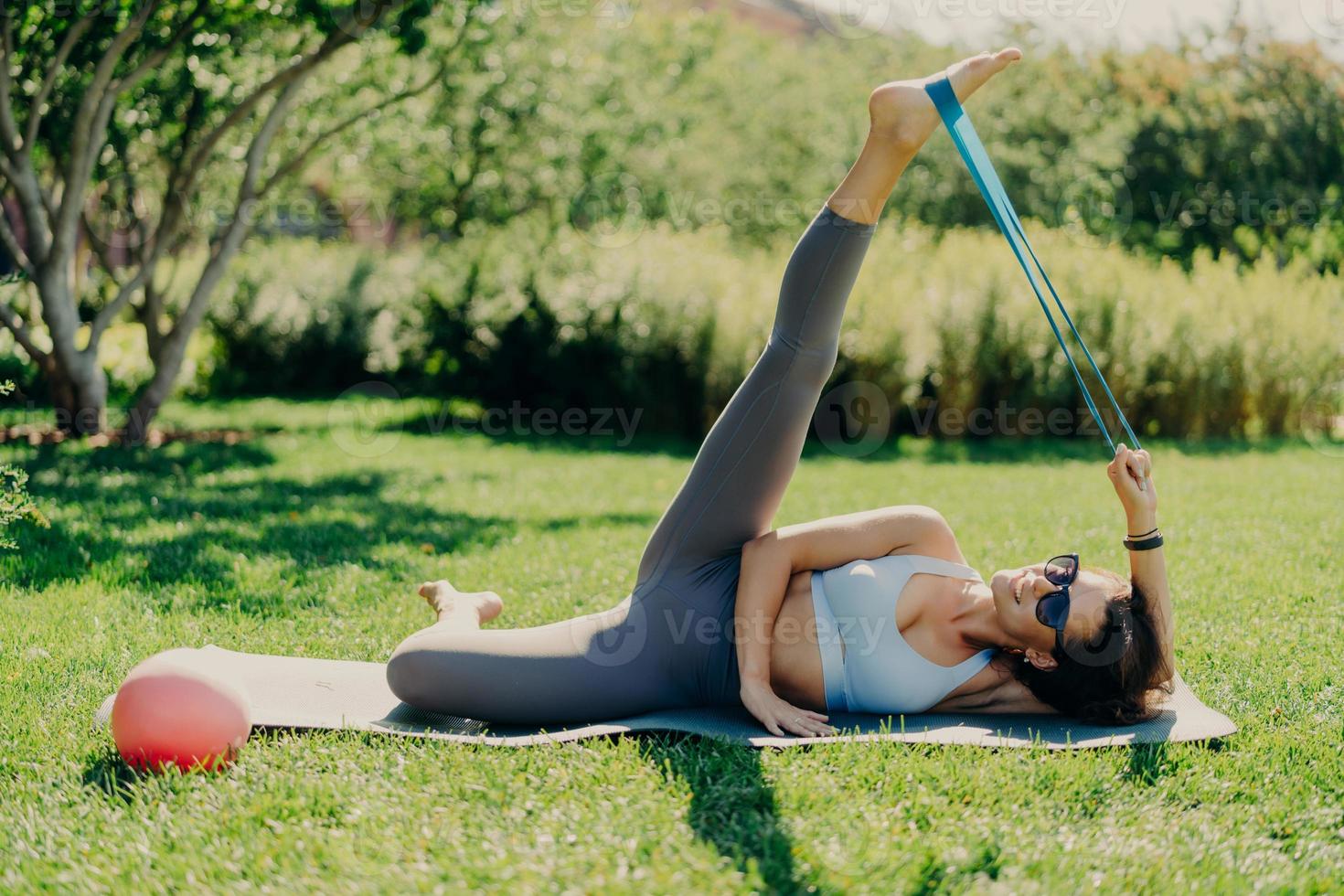 la mujer activa hace ejercicio al aire libre. mujer morena delgada estira las piernas con chicle deportivo vestido con ropa activa tumbada en la alfombra de fitness contra el fondo verde de la naturaleza tiene buena flexibilidad. haciendo ejercicio foto