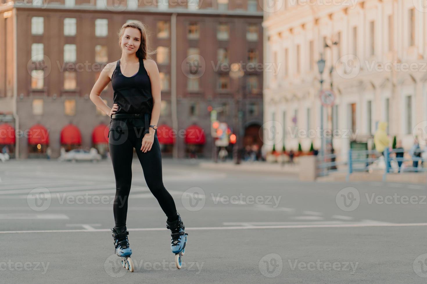 Sport and recreation concept. Horizontal shot of active woman practices rollerblading poses on blades keeps hand on waist blurred city background takes break. Rolleskating after work. Healthy life photo