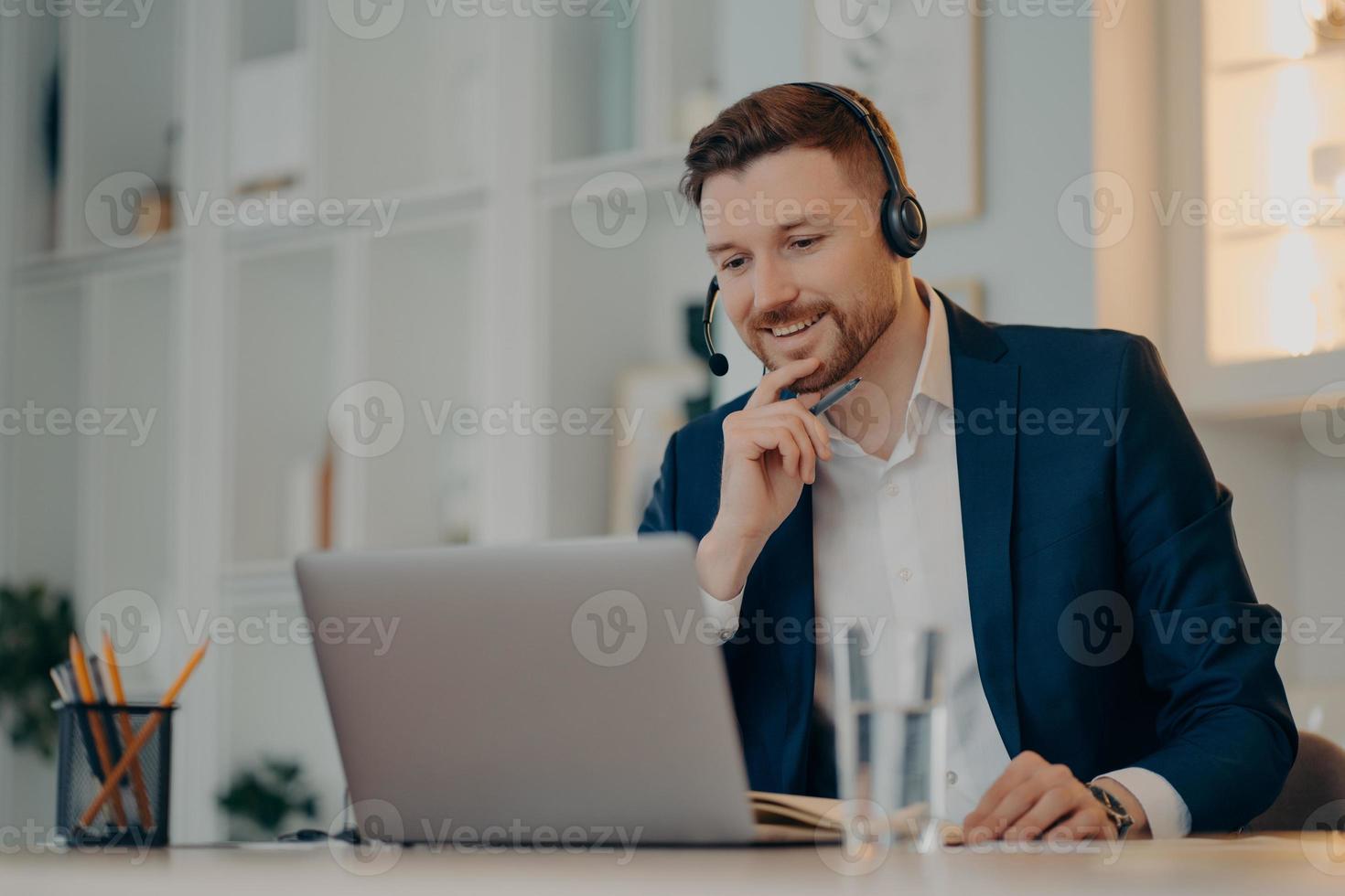 Professional male operator works in callcenter uses headset and laptop computer talks on video call or virtual webcam event wears formal clothes poses against cozy interior has online meeting photo