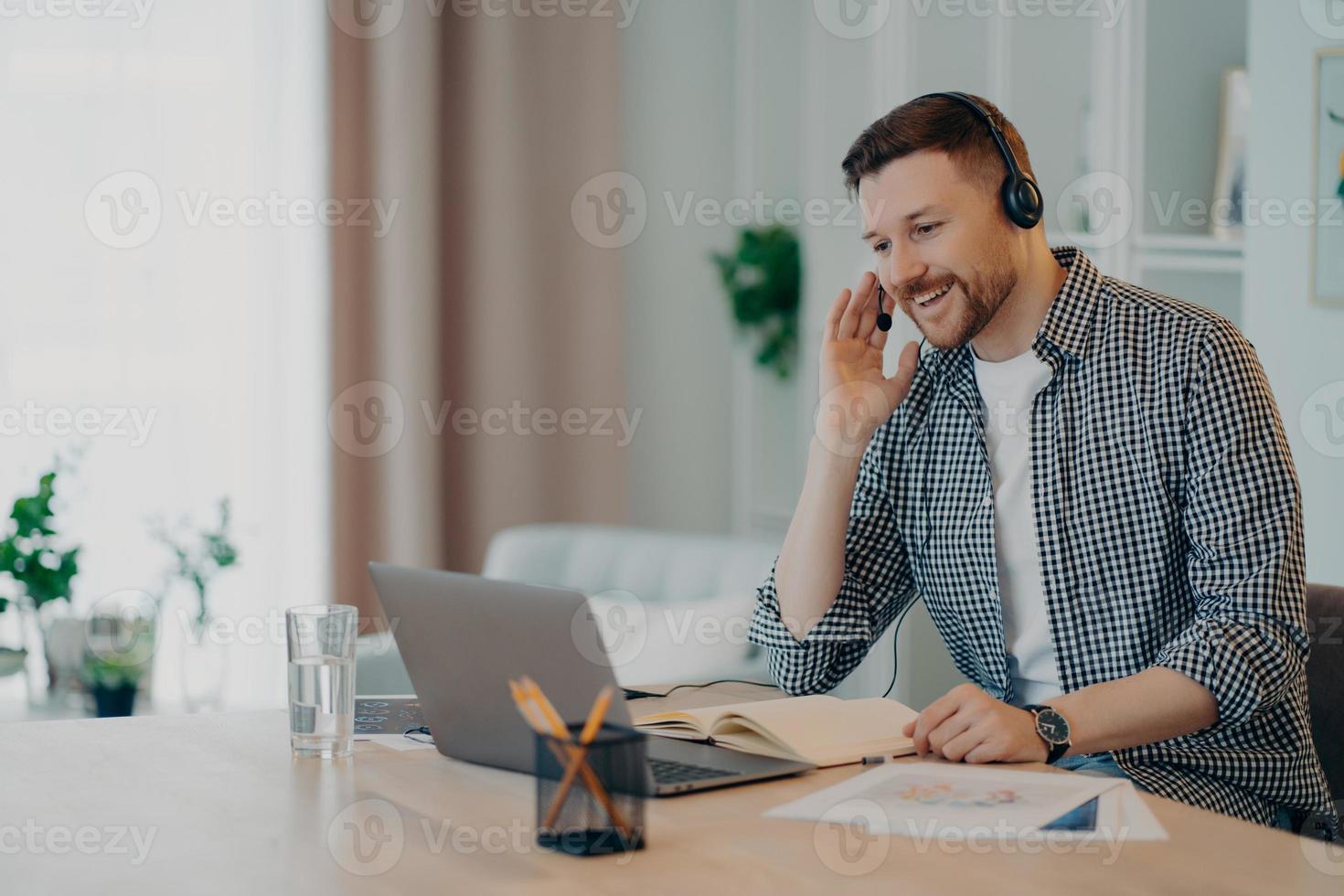 Horizontal shot of cheerful man participates in self improvement webinar wears checkered shirt communicates online by video call uses headset and laptop computer studies online poses at desktop photo