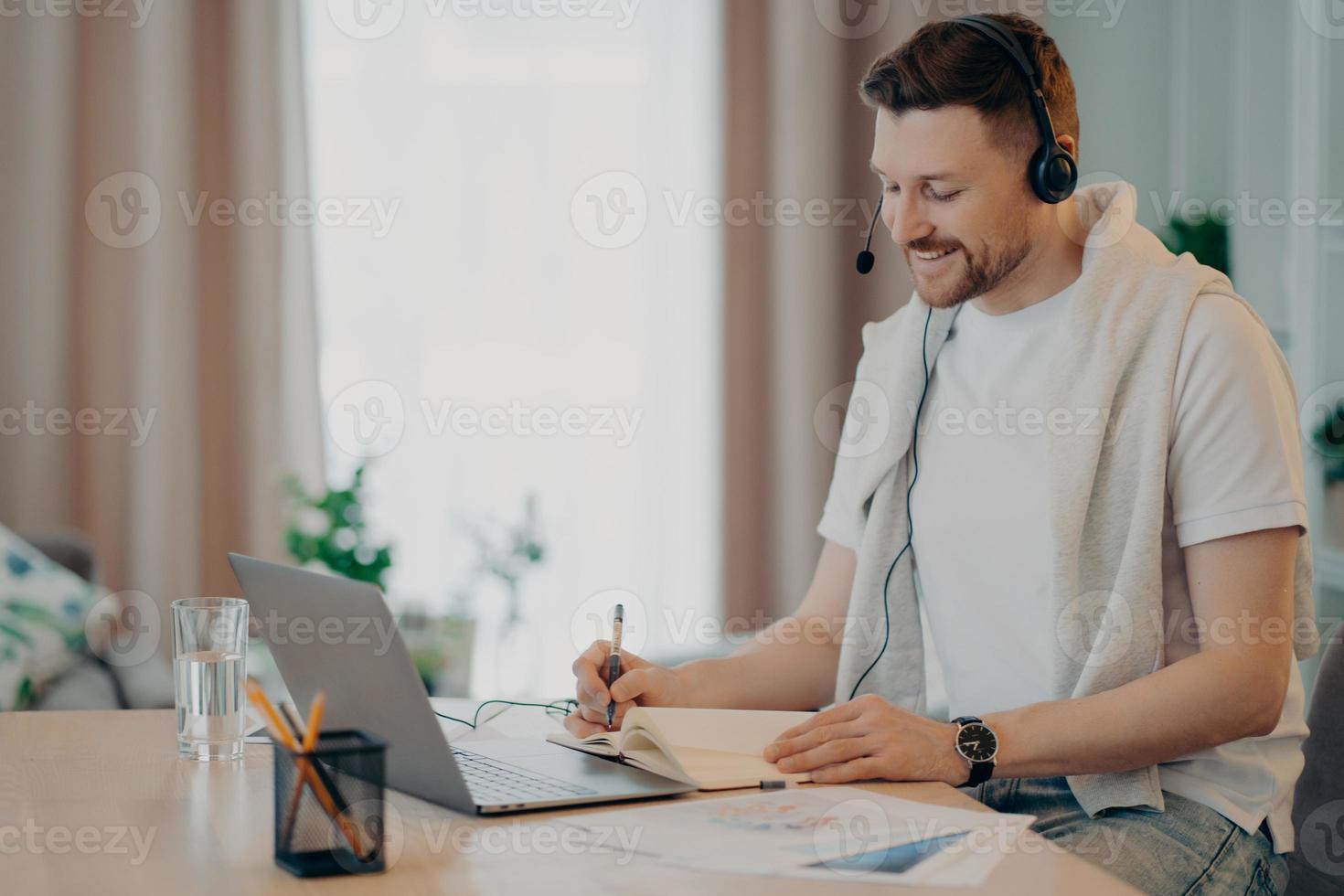 Happy young man sitting at the table at home and making notes in note book photo