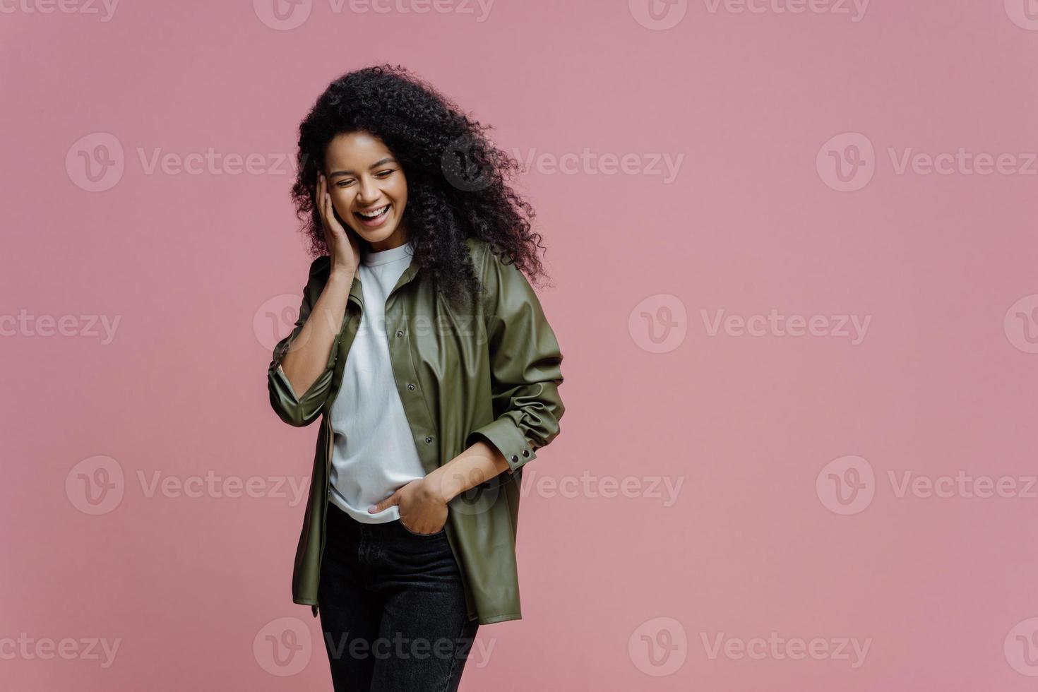 una foto horizontal de una mujer afroamericana feliz mantiene la mano en el bolsillo, se ríe mientras escucha una anécdota graciosa, pasa el tiempo libre en compañía alegre, se concentra, aislada en un fondo rosa, copia espacio