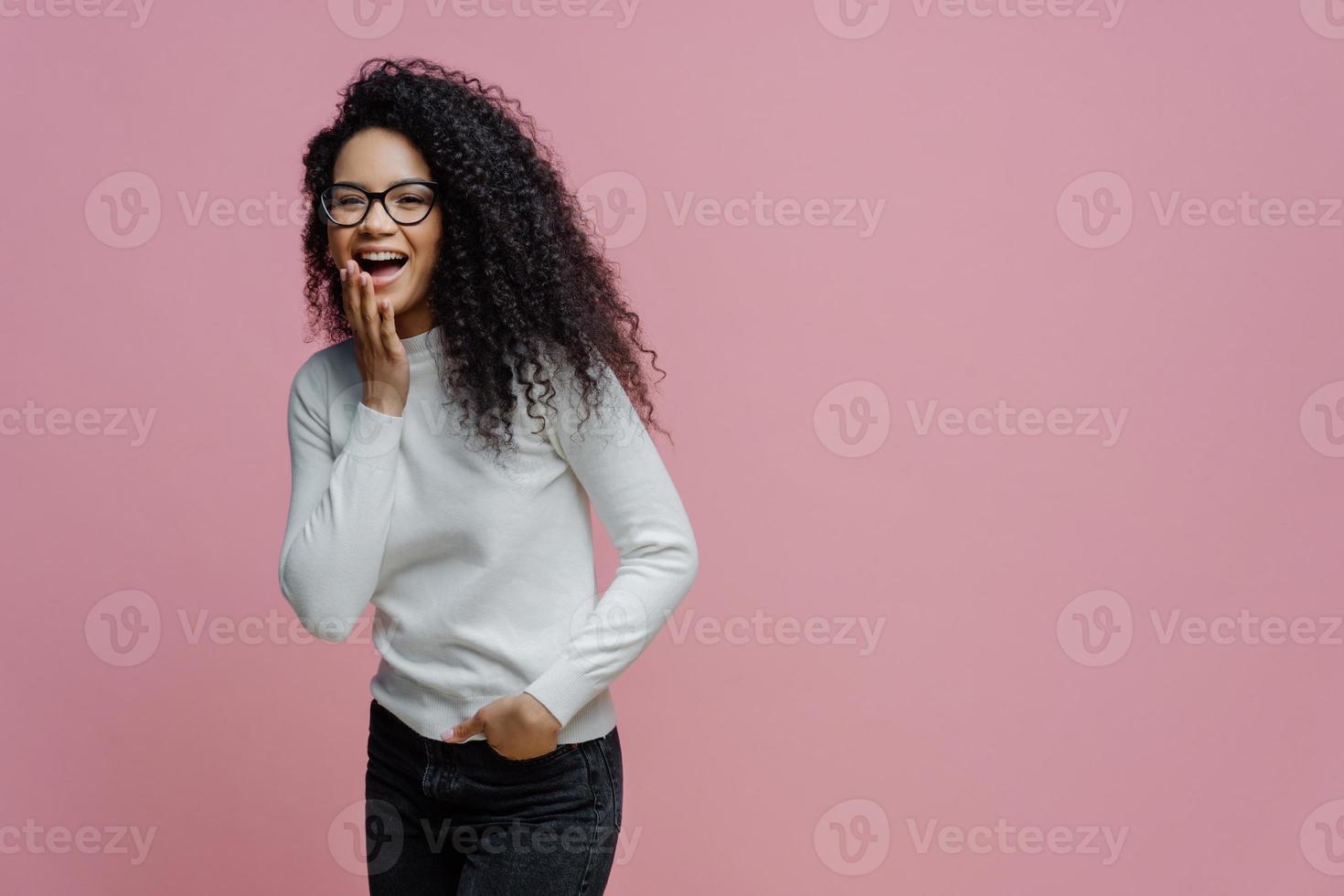 mujer afroamericana positiva se ríe de algo gracioso, cubre la boca abierta con la palma, no puede controlar sus emociones, mantiene la mano en el bolsillo de los jeans, usa poloneck blanco, aislada en un fondo rosado foto