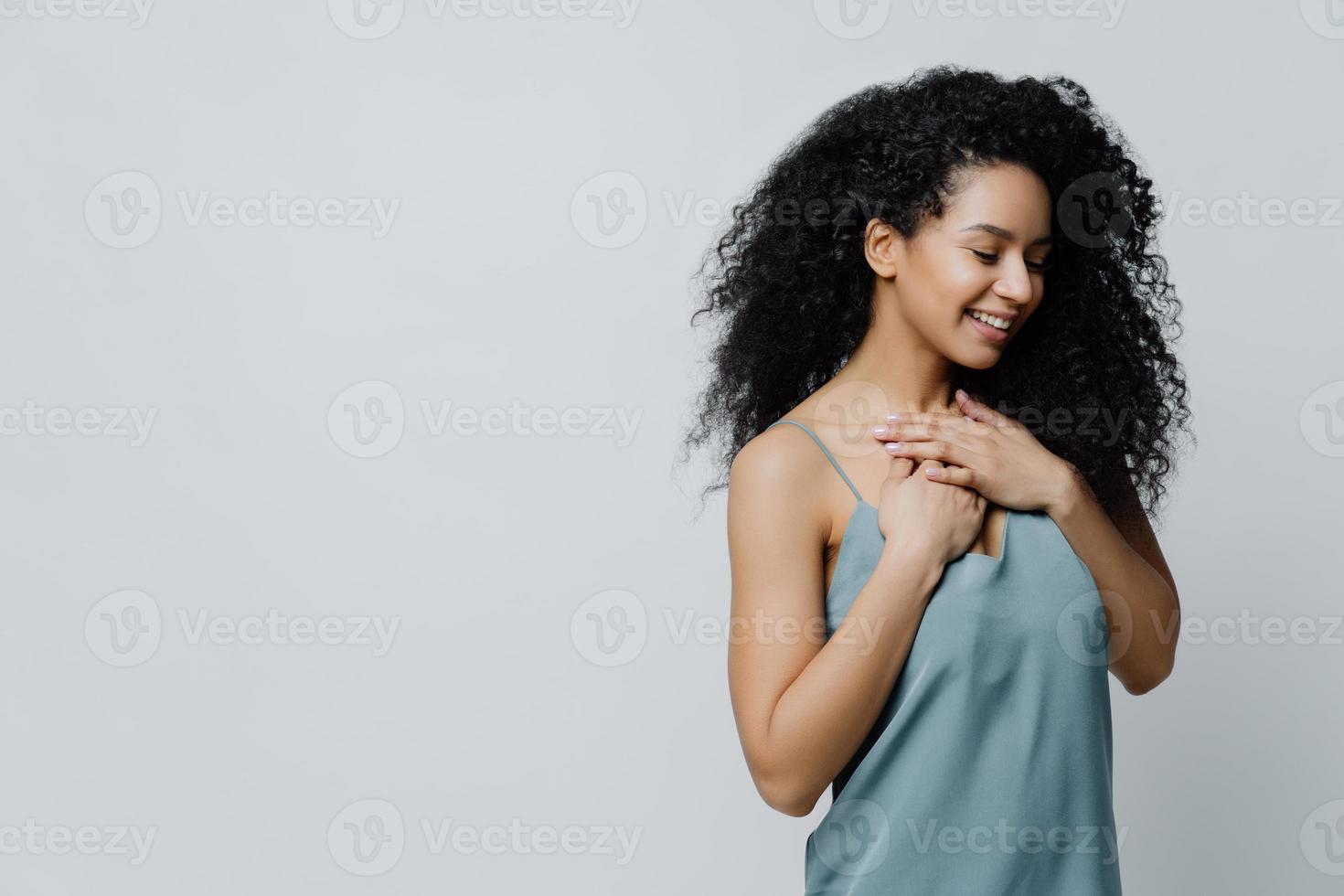 Profile of dreamy romantic ethnic woman with Afro haircut, dressed in nightwear, keeps hands on chest, recalls very pleasant moment in life smiles tenderly with closes eyes expresses truthful feelings photo