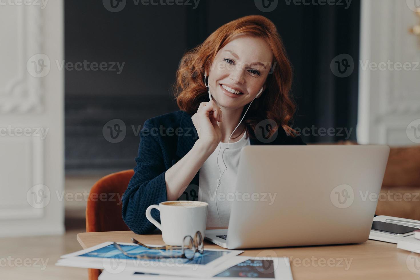 Freelance business woman with ginger hair sits in front of laptop computer, communicates with colleagues via video conference, sits at desktop, drinks coffee, has happy expression. Distance job photo
