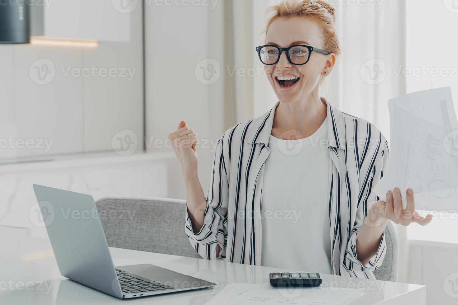 Overjoyed ginger woman holds papers works on project statistics celebrates finishing of work photo