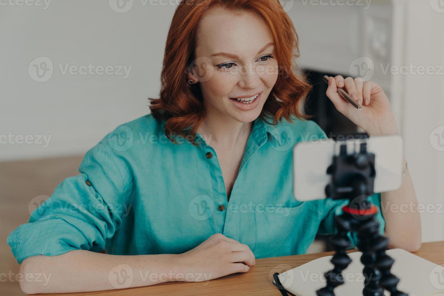 Young professional woman vlogger or blogger poses in front of smartphone camera, gives advice how to start own business, sits at desktop, holds pen. People, technology, lifestyle, blogging concept photo