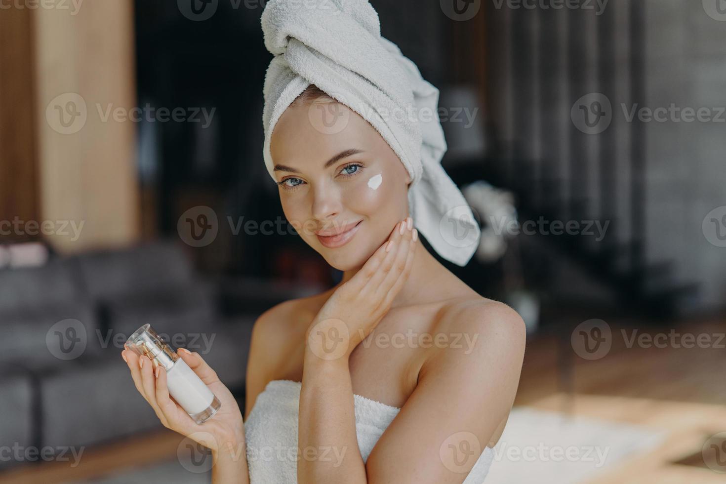 Beautiful young European woman applies face cream, hydrates skin, wears minimal makeup, wears wrapped towel on head, poses against home interior, undergoes facial treatment. Anti aginng procedure photo