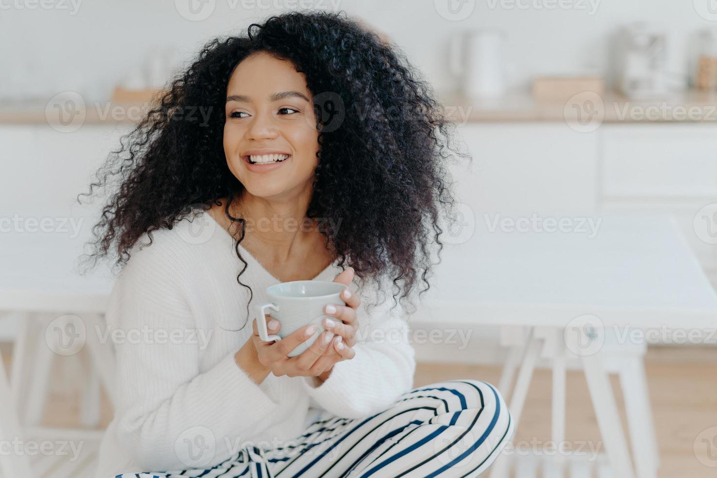 la foto de una linda mujer alegre con el pelo rizado tupido mira hacia otro lado con una sonrisa, sostiene una taza de café, usa ropa informal, disfruta de una mañana tranquila y acogedora, saborea el sabor. afro yacía en la cocina durante el día libre