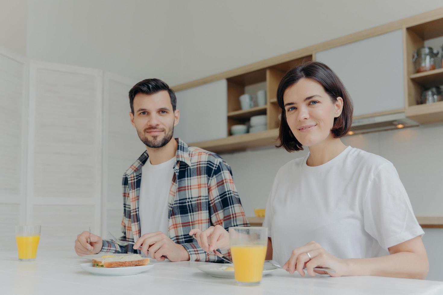 concepto de familia y nutrición. esposo y esposa miran alegremente a la cámara, disfrutan de un delicioso desayuno, beben jugo recién hecho, discuten sus planes familiares, posan en una cocina moderna, ambiente acogedor foto