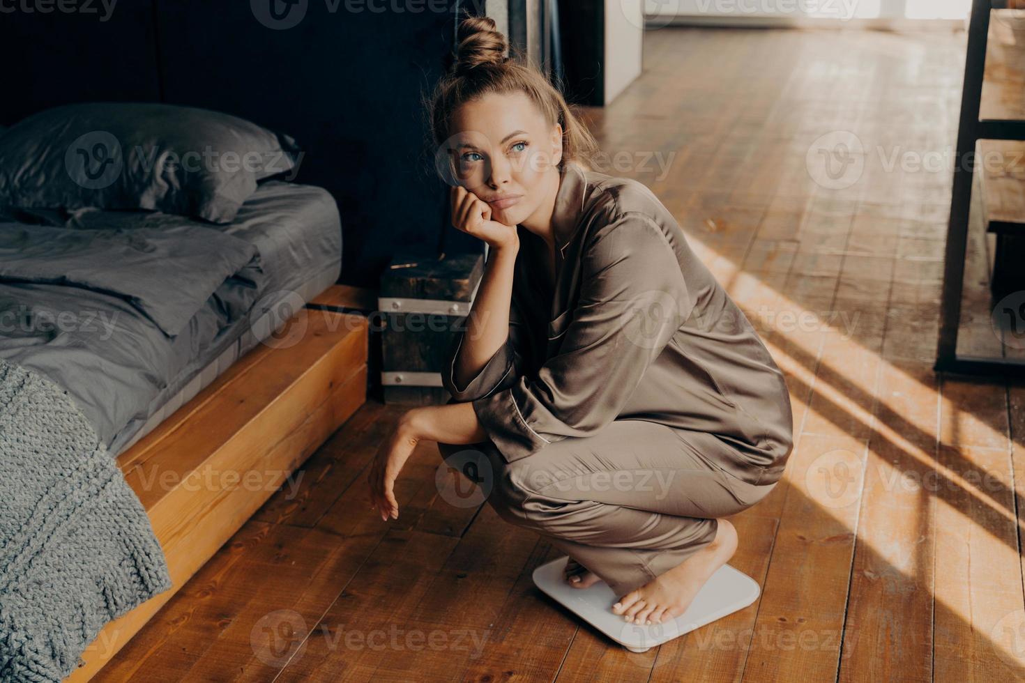 joven mujer molesta en pijama satinado midiendo su peso en casa foto